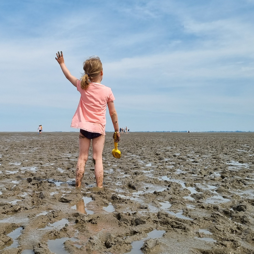 Kind winkt mit Schaufel in der Hand Menschen weiter weg im Watt zu
